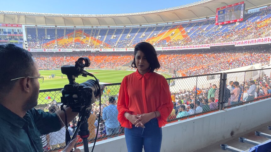 A woman in a bright red top stands in front of a camera inside a stadium