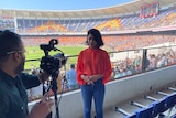 A woman in a bright red top stands in front of a camera inside a stadium