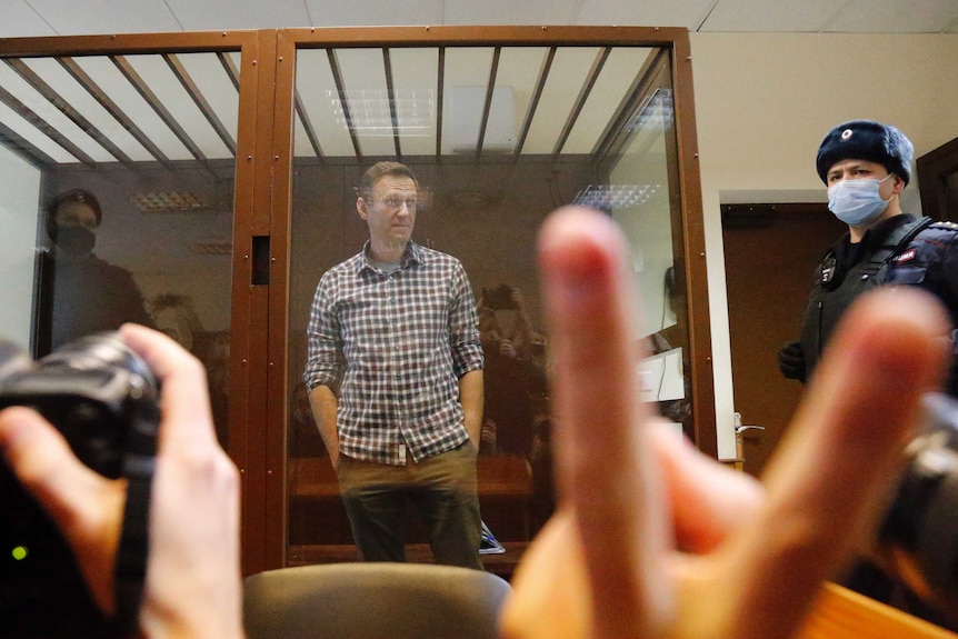 A man in a court dock stands with hands in his pockets as guards look on and media take photographs.