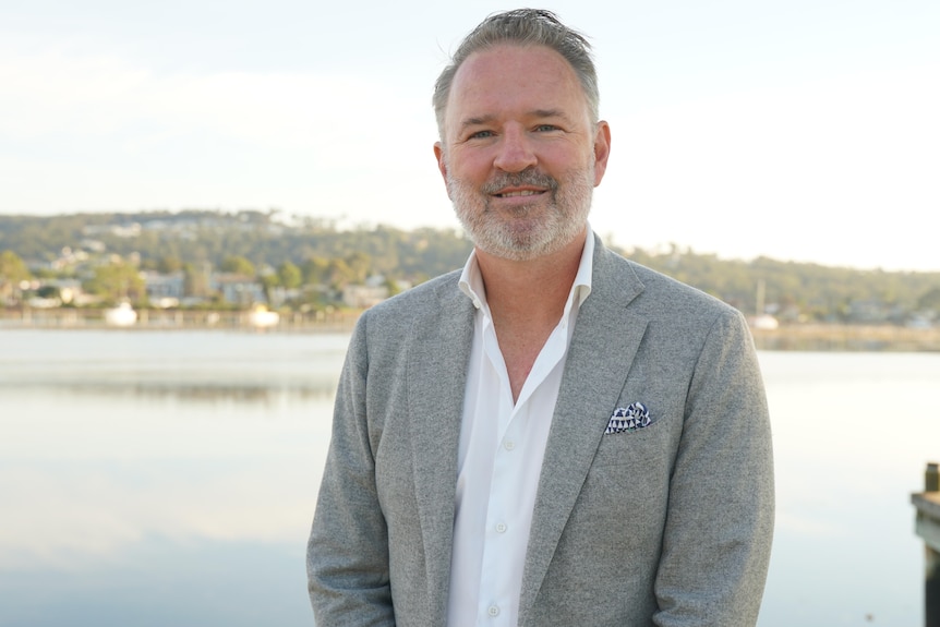 A man, wearing a white shirt and a grey blazer, stands near the ocean.