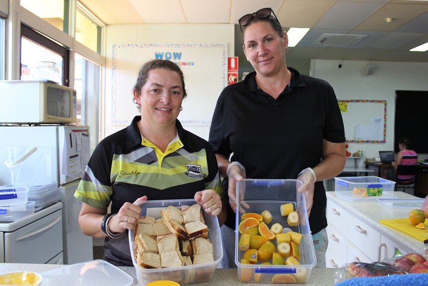 Two women in a school canteen show off tubs of sandwiches and fruit.