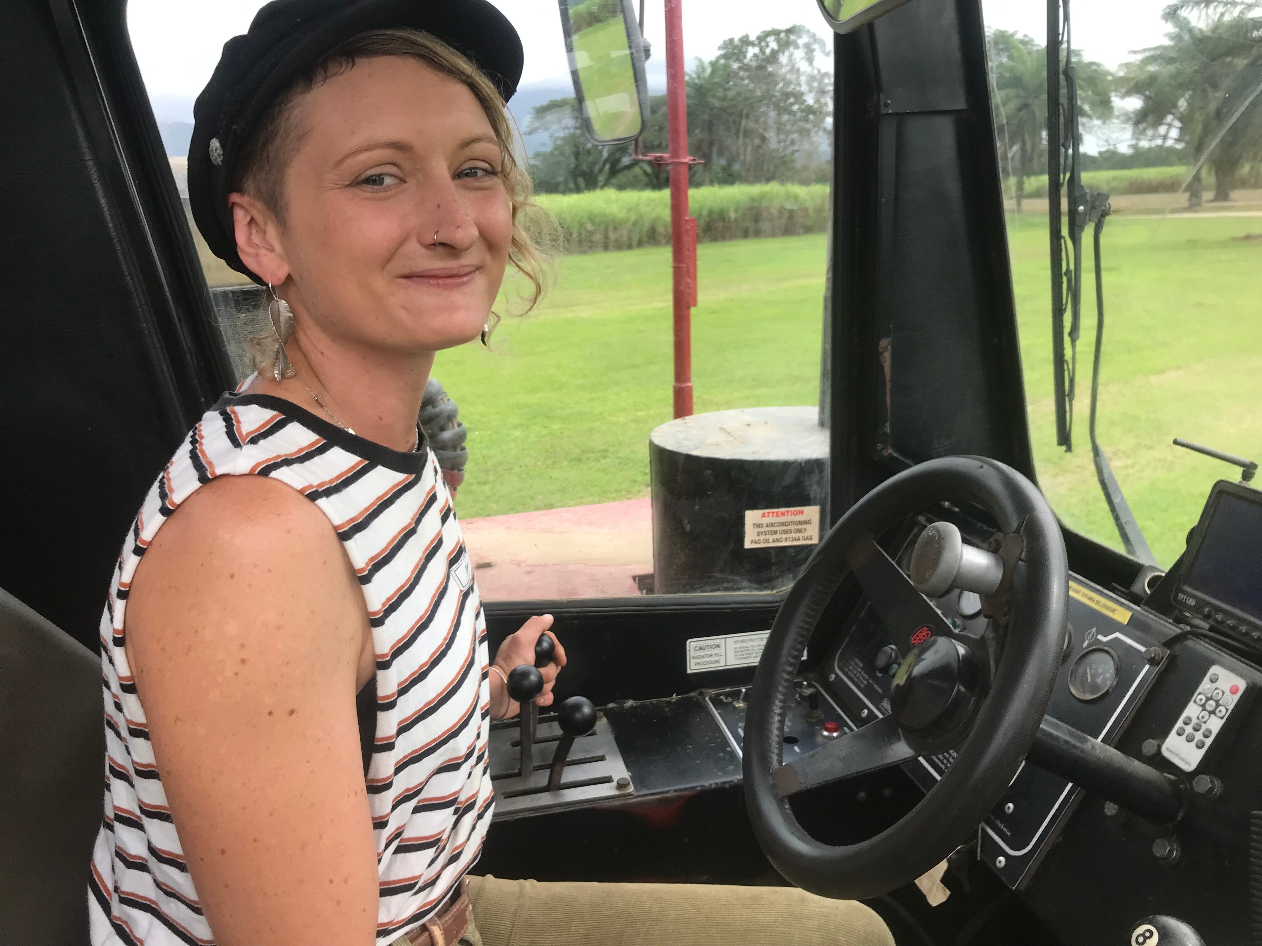 A young woman with short blond hair, a singlet and a black hat sits in the cab of a power-haul vehicle.
