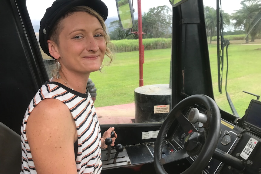 A young woman with short blond hair, a singlet and a black hat sits in the cab of a power-haul vehicle.