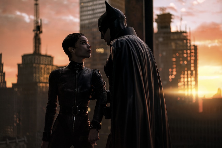 Woman with short dark hair wears black latex cat suit and stands intimately with man in black latex caped suit and mask.