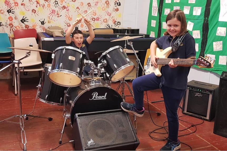 A boy sits behind a drum kit while a girl stands with one foot on a speaker while holding a guitar.