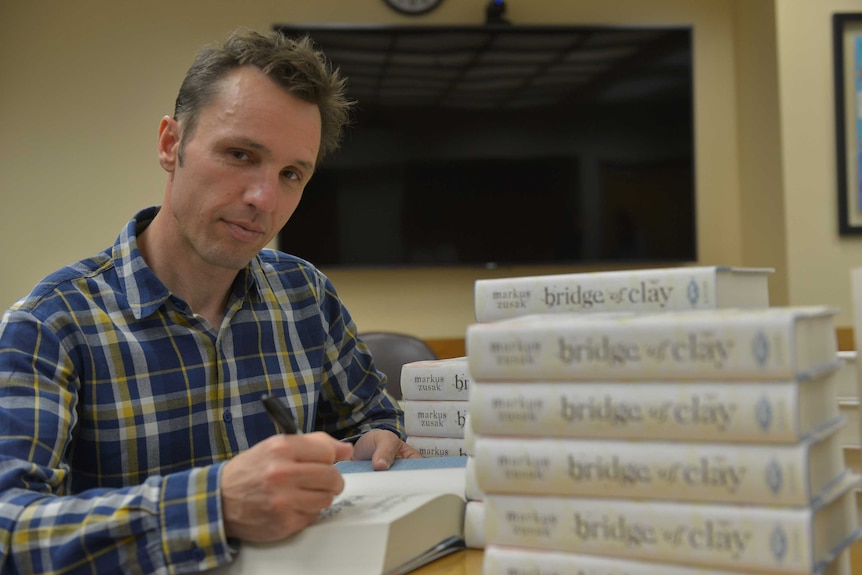 A young man stares into the camera while signing a book, a stack of books in front of him