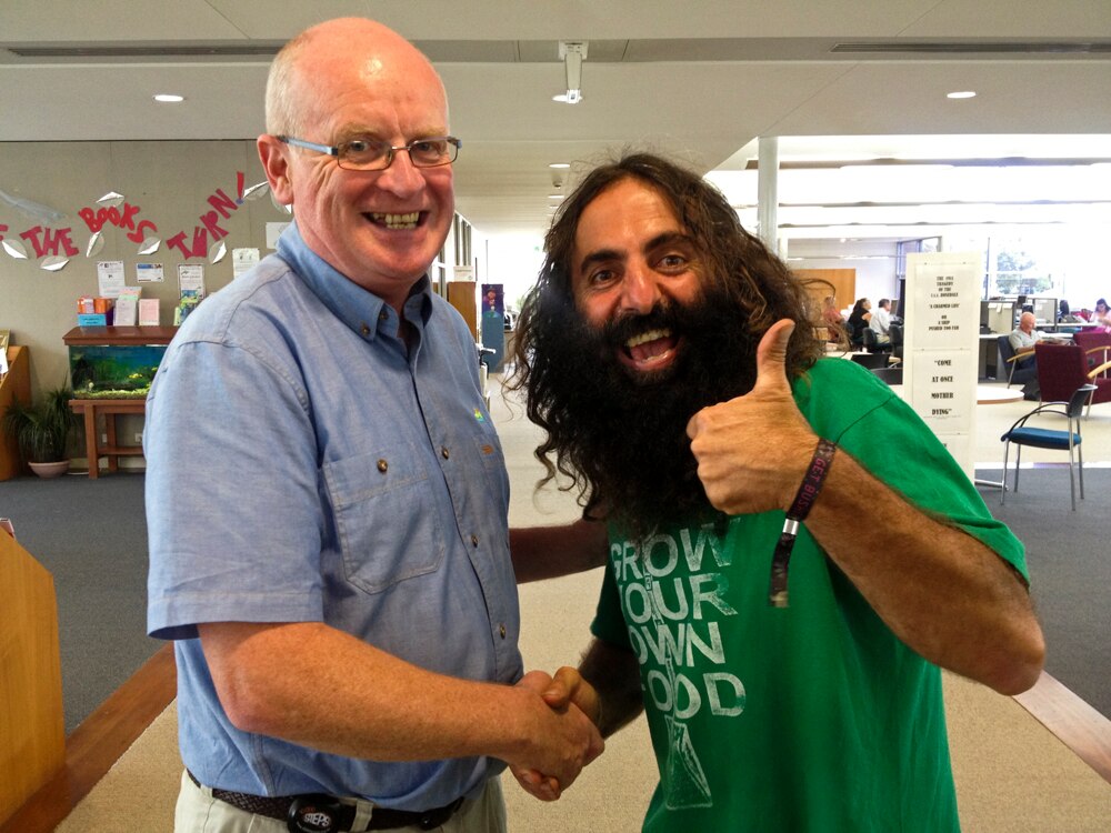 A Greek man with a bushy beard gives the thumbs up as he poses with a bald man wearing glasses in a library