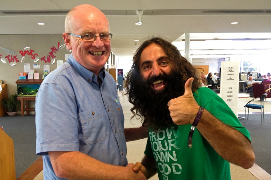 A Greek man with a bushy beard gives the thumbs up as he poses with a bald man wearing glasses in a library