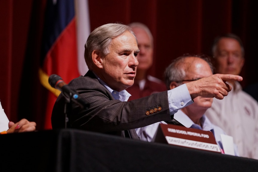 A middle-aged white man in a checked suit points while seated behind a table at a news conference.
