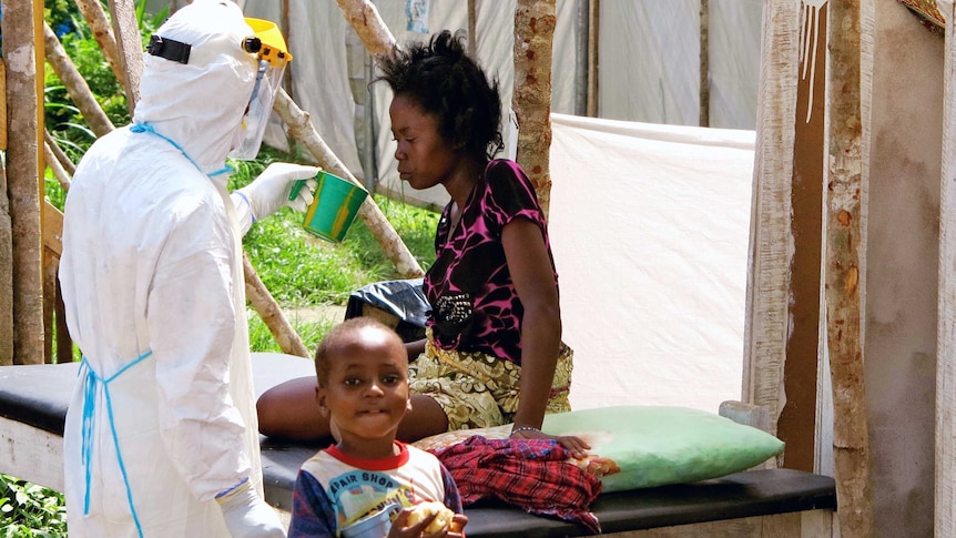 A health worker, wearing head-to-toe protective gear, offers water to a woman with Ebola.