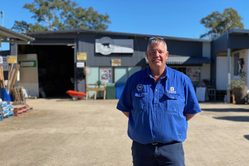 A man in a blue shirt standing outside a shed smiling.