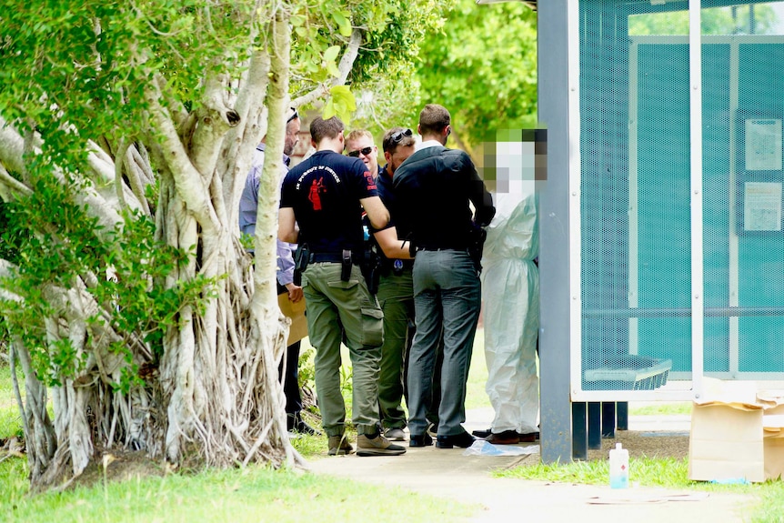 A group of detectives surround a man wearing a white forensic suit near a bus stop.