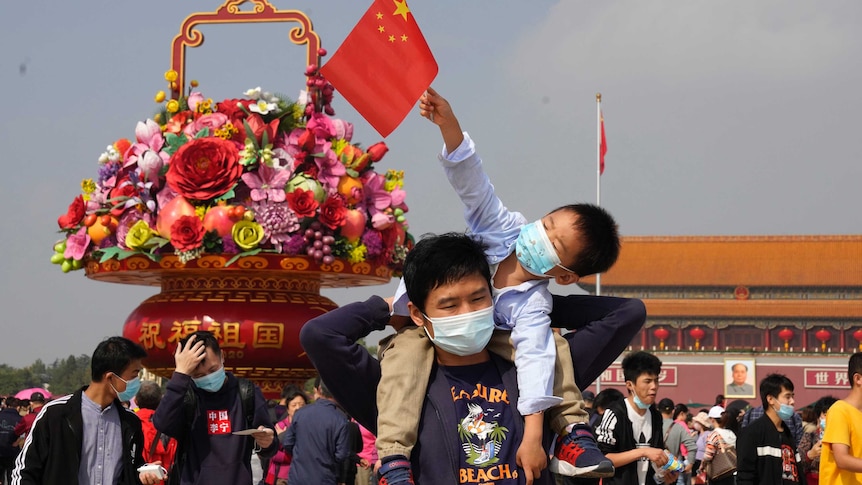 Tourists wearing masks to protect from the coronavirus stand near a floral decoration.