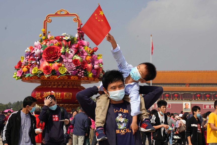 Tourists wearing masks to protect from the coronavirus stand near a floral decoration.