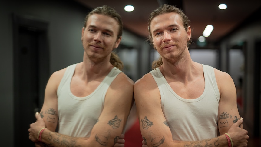 Young man wearing white singlet leans against mirror, in hallway.