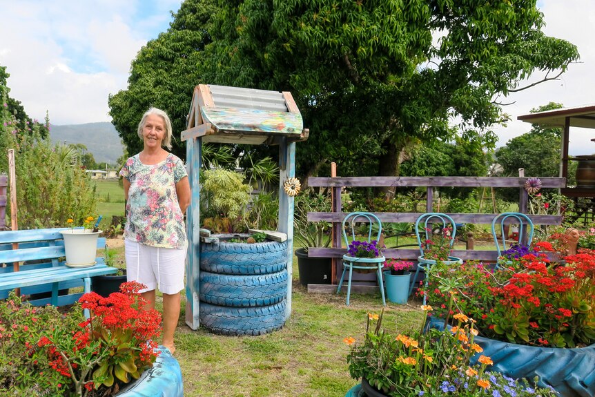 Robyn Smith dressed in  blouse with flowers on it stands in a vibrant colourful garden, a makeshift well stands behind her.