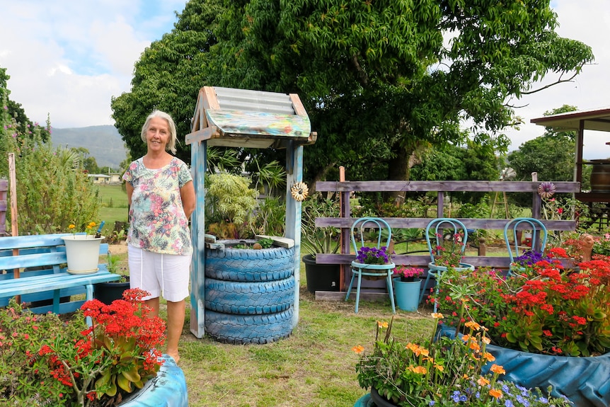 Robyn Smith dressed in  blouse with flowers on it stands in a vibrant colourful garden, a makeshift well stands behind her.