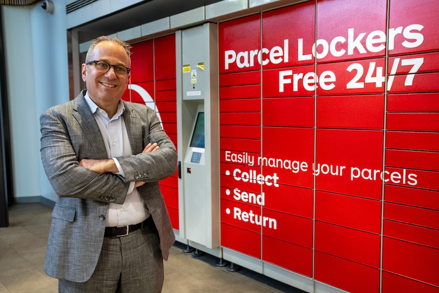 A man with a great suit, grey hair and glasses stands in from of a red wall with the words 'parcel lockers' on it. 