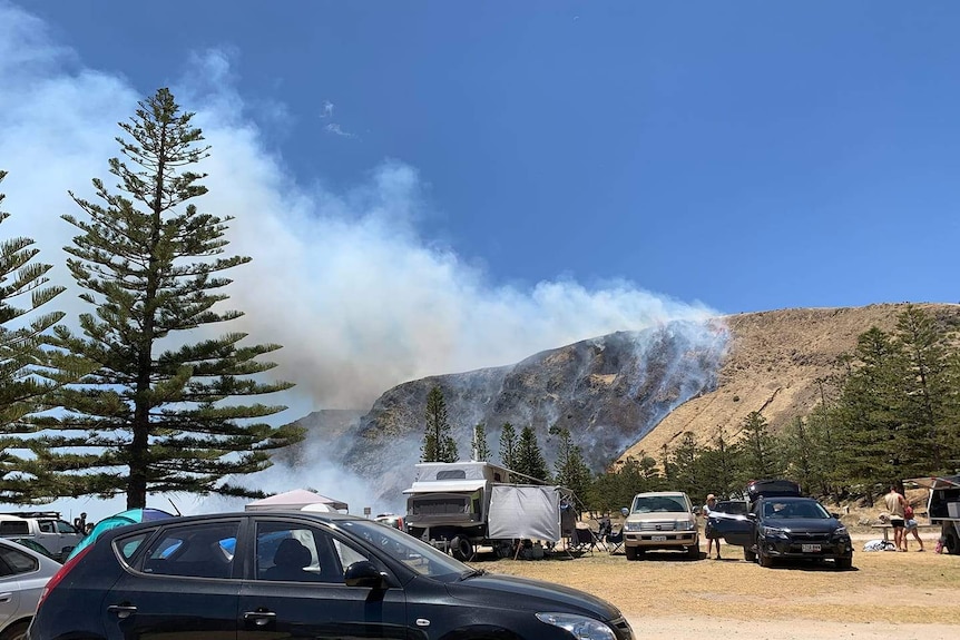 Smoke billows from a fire burning on a hillside with a caravan park in the foreground.