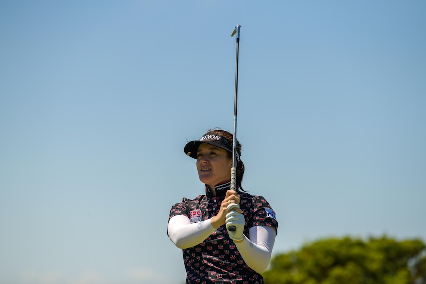 A woman in a hat holds a golf club in the air as she watches something in the distance