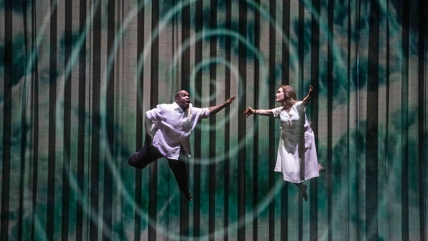 A Black male opera singer and a white female singer are suspended in mid-air above a stage against a swirling backdrop.
