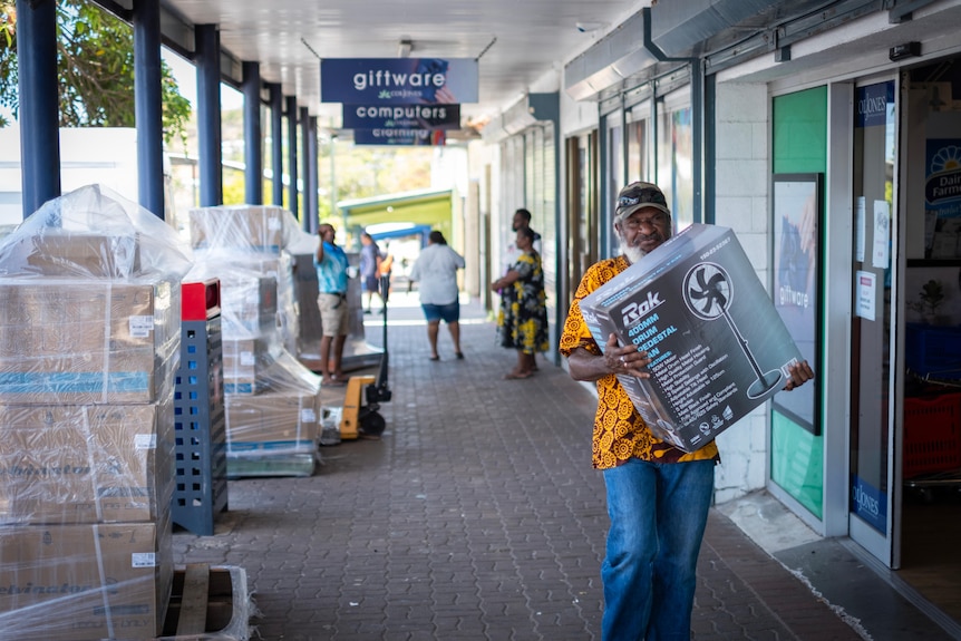 A man carrying a boxed pedestal fan down a shopping strip with pallets of whitegoods to the side of the footpath.