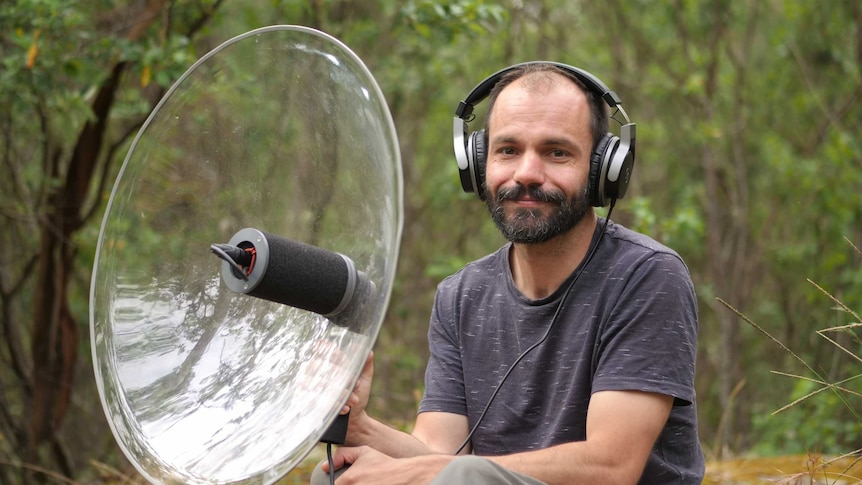 A man wearing headphones holds a large parabolic dish in a forest.
