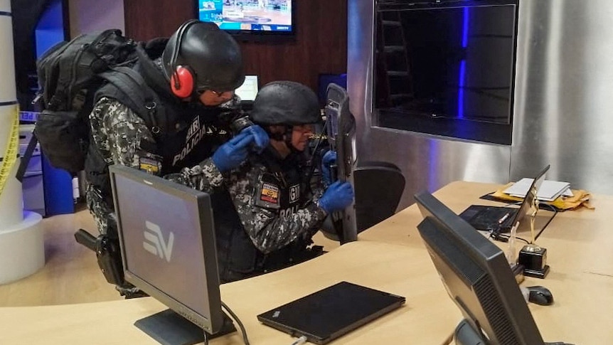 Two men wearing military fatigues and body armour inspect a desk with computer screen and a laptop