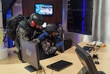Two men wearing military fatigues and body armour inspect a desk with computer screen and a laptop