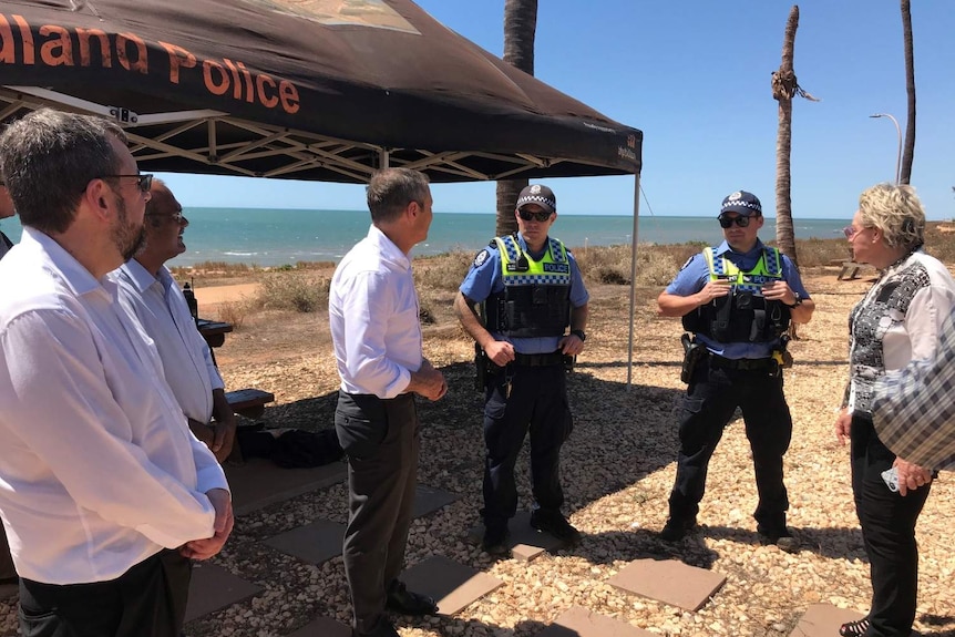 Roger Cook stands in the centre of a group of people, with two uniformed police officers, on the foreshore of a beach.