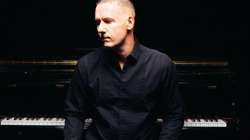 man in black shirt sitting with hands on knees with a piano in background