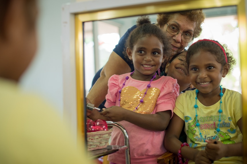 Three young indigenous girls look at their reflection in a mirror along with their kindergarten teacher.