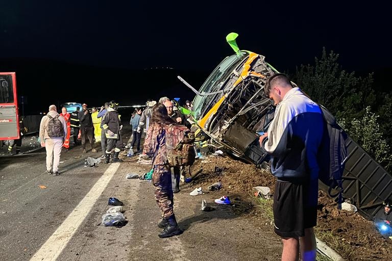 Passengers gather around a bus, flipped on its side, in a ditch, after a crash at night off a highway.
