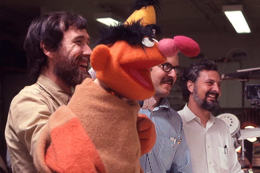 Three white and bearded men with dark hair look off camera smiling. Two are holding puppets and all are wearing button up shirts
