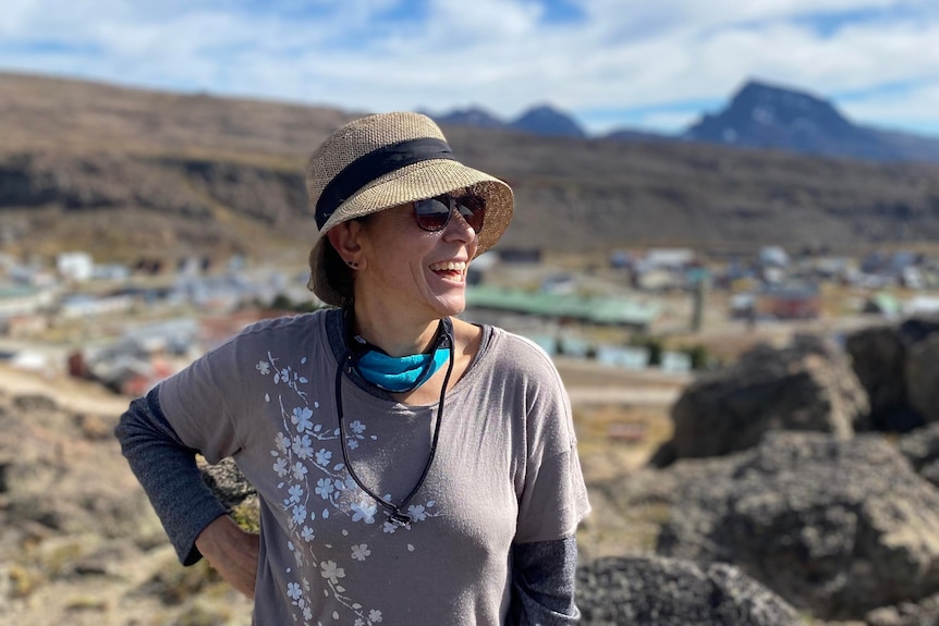 A smiling middle-aged woman in a hat and sunglasses overlooks a rocky landscape.