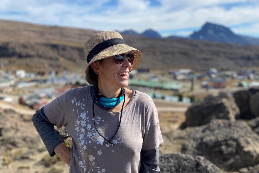 A smiling middle-aged woman in a hat and sunglasses looks out across a rocky landscape.