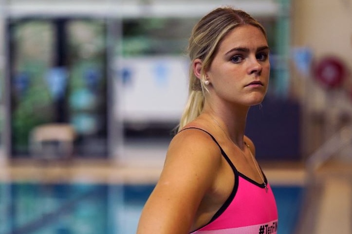 A young blonde woman stands beside a pool wearing a neon pink swimsuit