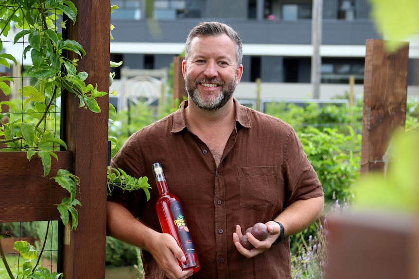 Ryan holds a bottle of passionfruit liqueur and some passionfruit while standing among passionfruit vines.