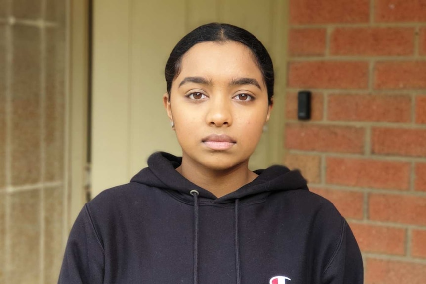 A young woman with her hair tied back wearing a black hoodie stands in front of her home.