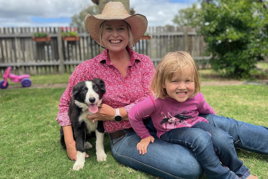 A mid shot of a woman seated with her 4yo daughter on her lap and a border collie puppie beside her