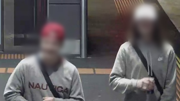 Two young males on CCTV footage, one wearing a red cap and one wearing a white cap. Both are wearing grey hoodies.