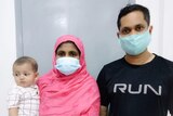 A woman in a pink headscarf and mask holds a baby in a checkered shirt next to a man wearing a mask and black tshirt