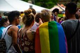 A group of young people, one draped in a rainbow pride flag, stand in a group of other youths at an outdoor venue.