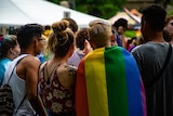 A group of young people, one draped in a rainbow pride flag, stand in a group of other youths at an outdoor venue.