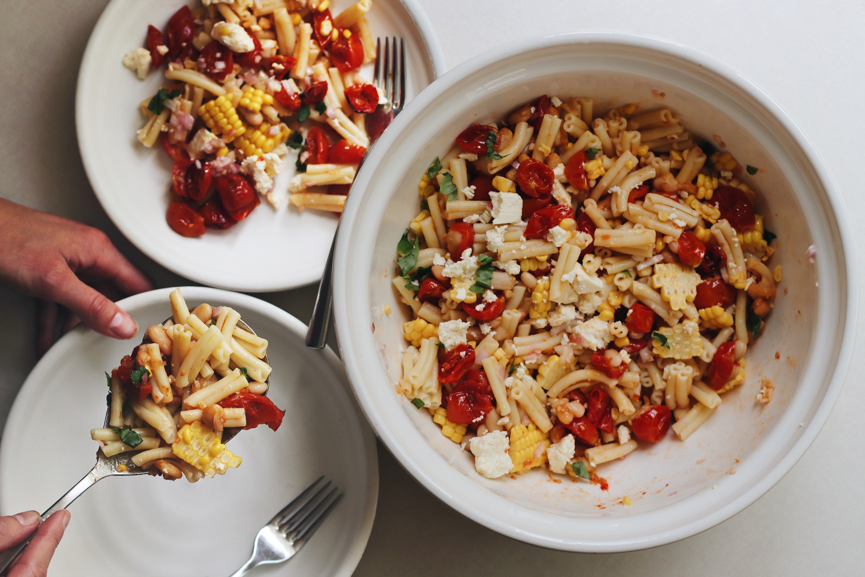 Close-up of hands serving pasta salad with corn, feta, tomatoes and basil on a plate, from a large bowl of pasta salad.