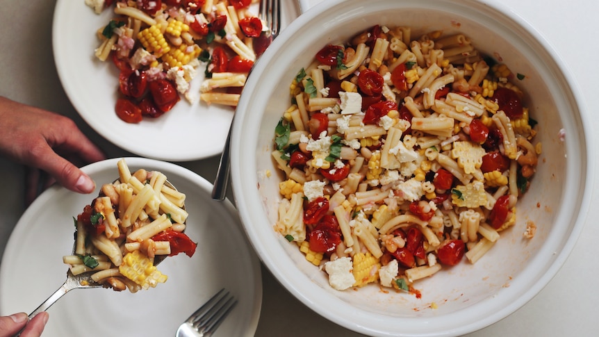 Close-up of hands serving pasta salad with corn, feta, tomatoes and basil on a plate, from a large bowl of pasta salad.