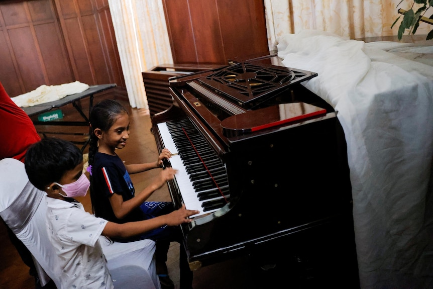 A young girl has her hand on the keys, as another young child sits with her on the piano stool