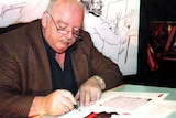 A man with glasses in a brown jacket writing at a desk