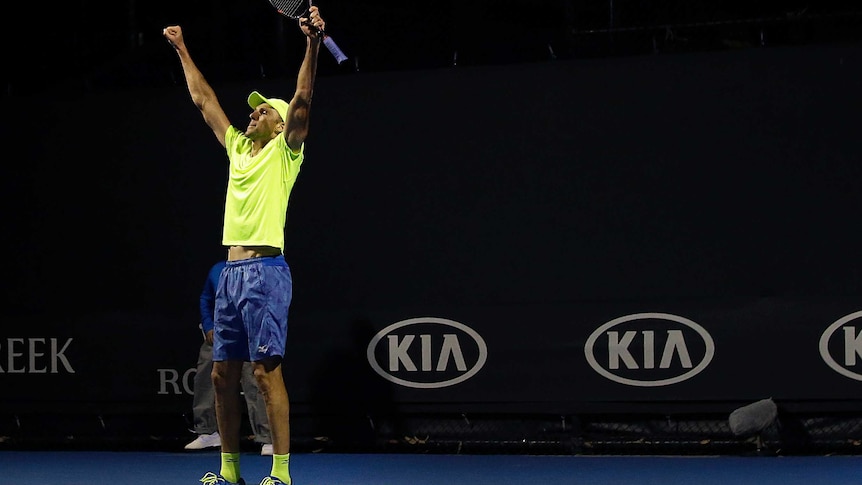 A tennis player in a fluoro yellow t-shirt raises his arms in celebration holding a tennis racquet.