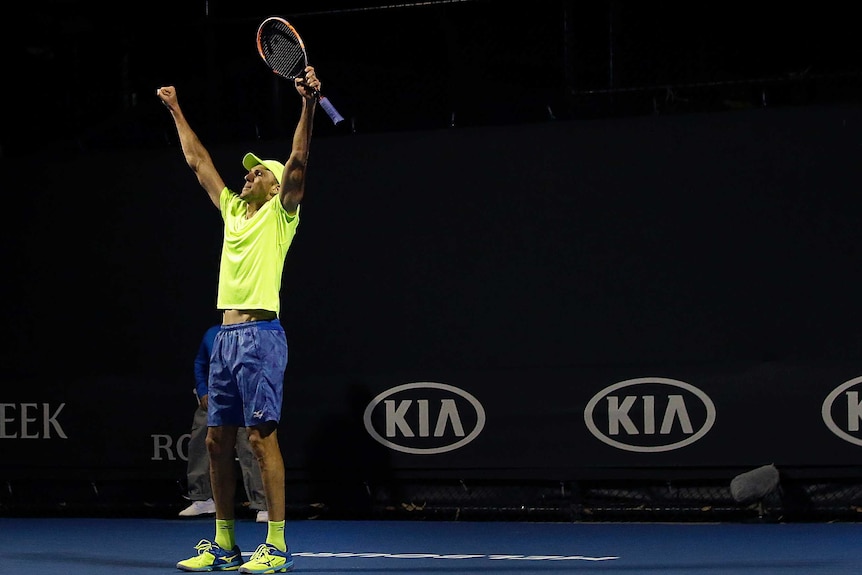 A tennis player in a fluoro yellow t-shirt raises his arms in celebration holding a tennis racquet.